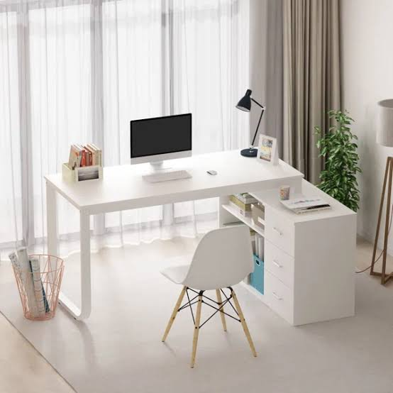 A white L-shaped office desk with drawers and open shelving, featuring a computer monitor, desk lamp, books, and office accessories. The bright minimalist setup demonstrates how an L-shaped desk can efficiently use corner space while providing a comfortable and productive workspace.