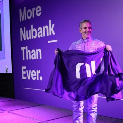 A promotional event for Nubank, featuring a person proudly displaying the Nubank logo on a purple banner. The image highlights brand promotion, financial innovation, digital banking, and Nubank’s growing presence in the financial services industry.