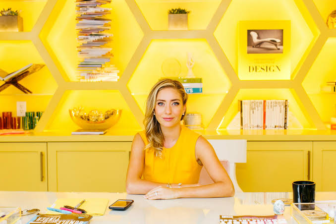 A modern professional woman sitting at a desk in a bright yellow office, surrounded by organized shelves, books, and stylish hexagonal wall décor. The image represents creativity, professionalism, interior design, modern business, and a vibrant workspace. It is suitable for articles or websites related to design, creative agencies, branding, architecture, business, or office interiors.
