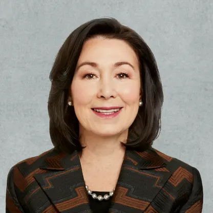 A woman with shoulder-length dark hair is smiling warmly at the camera. She is wearing a dark blazer with a brown geometric pattern, a black top, pearl earrings, and a pearl necklace. The simple gray background creates a clean and professional appearance.