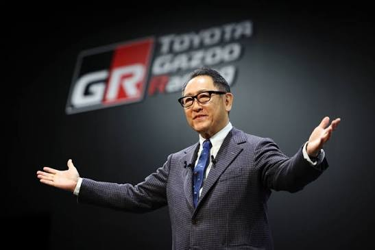 The image depicts a business executive presenting at an automotive industry event, likely related to Toyota Gazoo Racing. It communicates leadership, innovation, brand identity, and corporate communication.