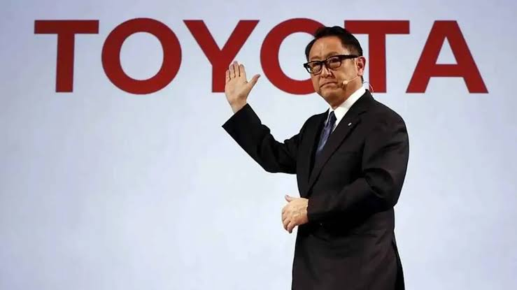 The image depicts a corporate executive speaking at a Toyota press conference or public presentation. It highlights corporate leadership, brand representation, and public communication in the automotive industry.