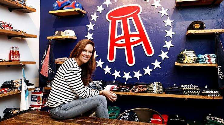 The image shows a woman associated with a sports media or lifestyle brand, posing inside a branded merchandise store. It highlights entrepreneurship, brand identity, and sports-related retail culture.
