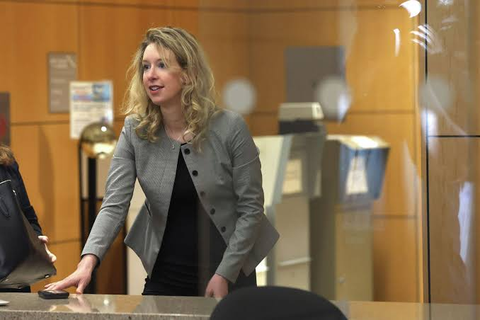 A woman wearing a grey blazer over a black top is standing behind a reception counter in a modern office or bank. She is leaning forward and interacting with a customer, possibly providing information, handling documents, or assisting with a financial transaction.